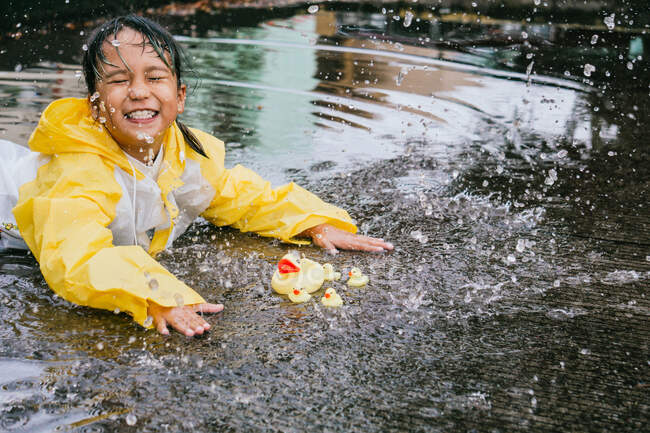 Cheerful Asian child with closed eyes in raincoat playing in puddle against toy ducks in rainy weather — Stock Photo
