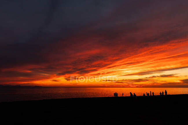 Vue panoramique de silhouettes touristiques admirant l'océan sans fin depuis le rivage sous un ciel nuageux avec un soleil brillant au crépuscule — Photo de stock