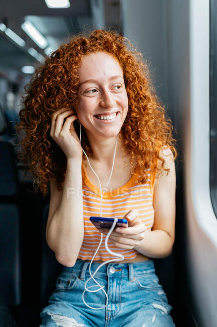 Candid young woman with curly red hair and cellphone listening to song from earphones while looking away on train — Stock Photo