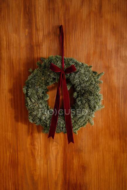 Grinalda conífera decorativa com arco vermelho pendurado na parede de madeira no quarto durante o Natal — Fotografia de Stock