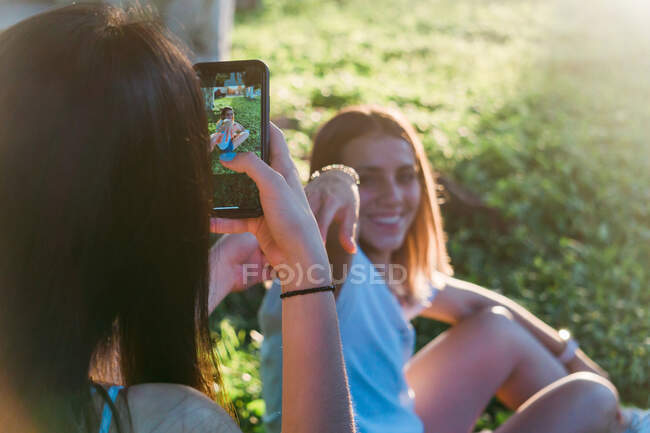 Crop unrecognizable female teenager taking photo of smiling best friend on cellphone while spending time on meadow in back lit — Stock Photo