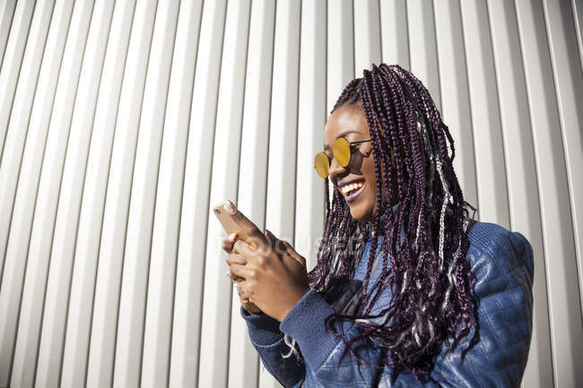 Cheerful stylish young African American female with Afro braids wearing trendy jacket and sunglasses browsing social networks on mobile phone while standing near wall of urban building — Stock Photo