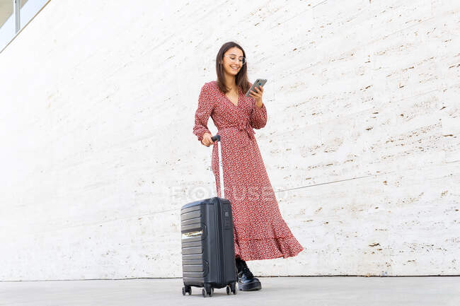 Позитивная женщина в длинном красном платье ходит с багажом на улице к белой стене днем — стоковое фото