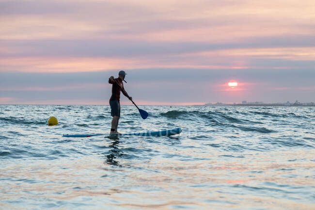 Vista lateral del surfista masculino en traje de neopreno y sombrero en tabla de paddle surfeando en la orilla del mar durante la puesta del sol - foto de stock
