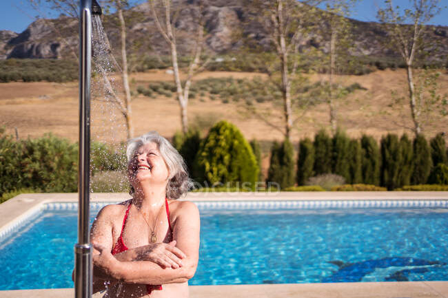 Mulher sênior alegre em biquíni desfrutando de salpicos de chuveiro perto da piscina com água clara transparente — Fotografia de Stock