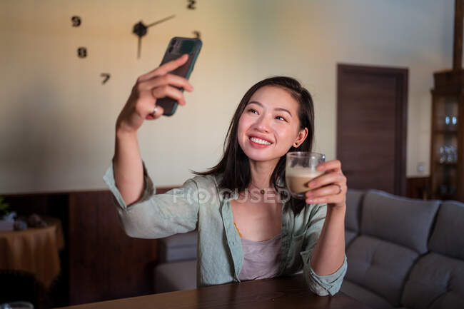 Content young ethnic female taking self portrait with coffee on cellphone while sitting on sofa with laptop at home — Stock Photo