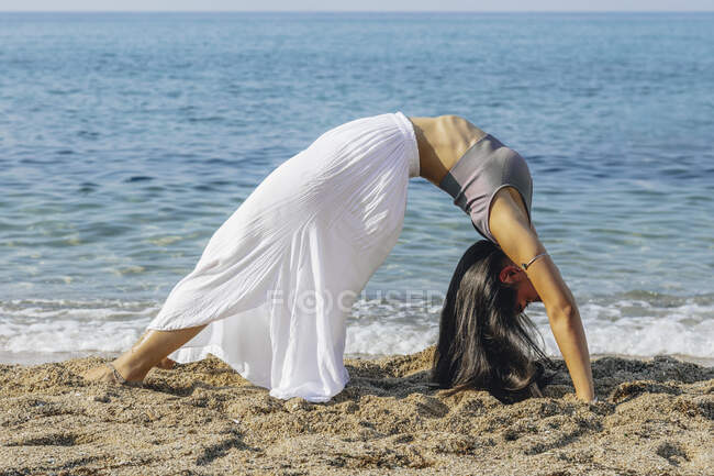 Vista lateral de la hembra de pie en Urdhva Dhanurasana posan durante la práctica de yoga en la costa arenosa contra el océano - foto de stock