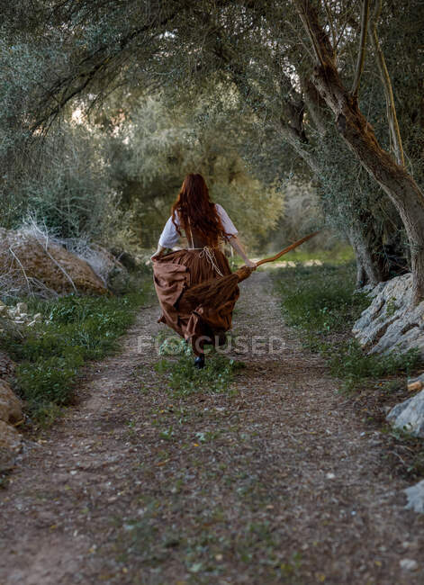 Обратный вид неузнаваемой ведьмы в одежде и с метлой, бегущей по тропинке в осеннем лесу — стоковое фото