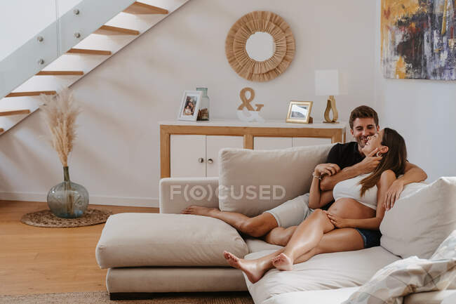 Cheerful man embracing belly of expectant female beloved while resting on couch in living room — Stock Photo