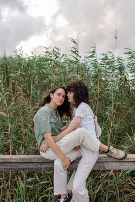 Side view of charming young homosexual girlfriends spending time on fence under cloudy sky in evening countryside — Stock Photo