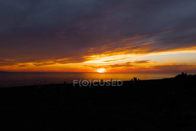 Scenic view of tourist silhouettes admiring endless ocean from shore under cloudy sky with shiny sun at dusk — Stock Photo