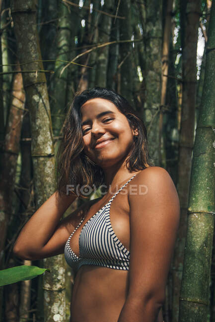 Contenida joven viajera en traje de baño con sombra en la cara mirando hacia otro lado contra ramas de bambú - foto de stock