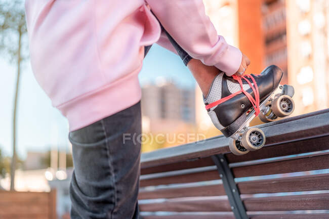 Vista lateral da fêmea jovem irreconhecível cortada vestindo capuz rosa claro e calças pretas amarrando patins de rolo perna inclinada na parte de trás do banco de madeira em parte — Fotografia de Stock