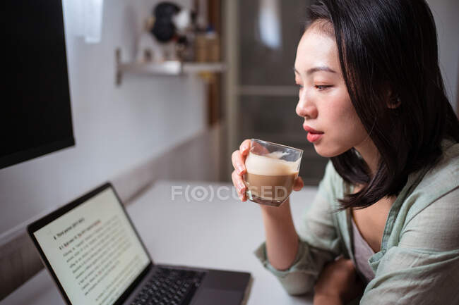 Vista lateral de la joven blogger étnica en el escritorio con netbook y café mirando a la cámara en la habitación de la casa - foto de stock