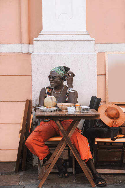 Прохолодний дорослий афроамериканець у сонячних окулярах і браслетах сидить за столом у міській кафетерії з кокосовим напоєм. — стокове фото