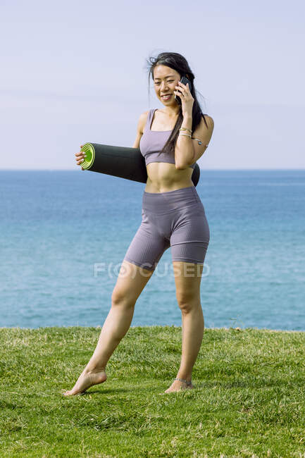 Sincero jovem étnico feminino em sportswear com tapete de ioga rolo falando no celular enquanto olha para baixo na praia do oceano — Fotografia de Stock