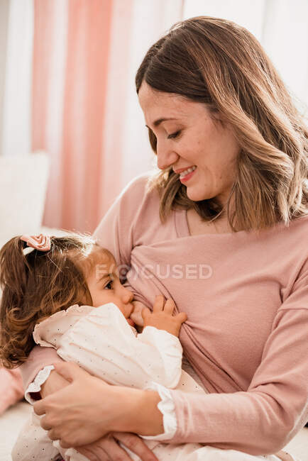 Positive adult mom feeding and tenderly embracing toddler daughter while sitting at home in daytime — Stock Photo