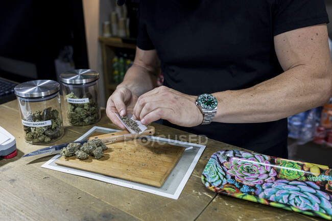 Crop macho anónimo en reloj de pulsera con hojas secas de marihuana molida en papel de cigarrillo sobre capullos de flores en tabla de cortar - foto de stock