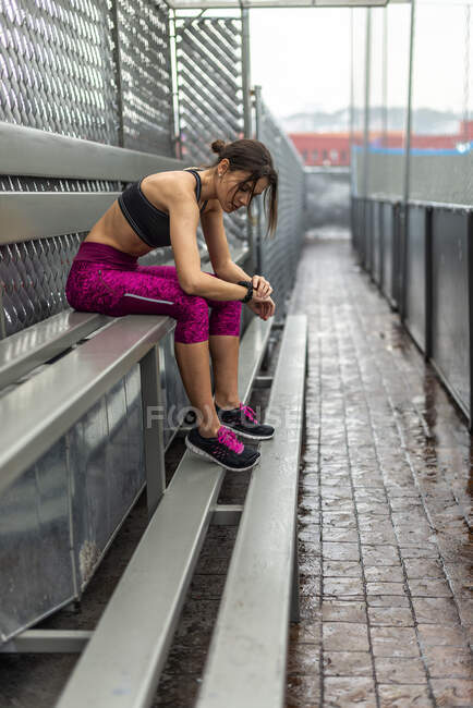 Vista laterale corpo pieno di atleta donna tonica in activewear seduto sulla panchina dopo l'allenamento all'aperto — Foto stock