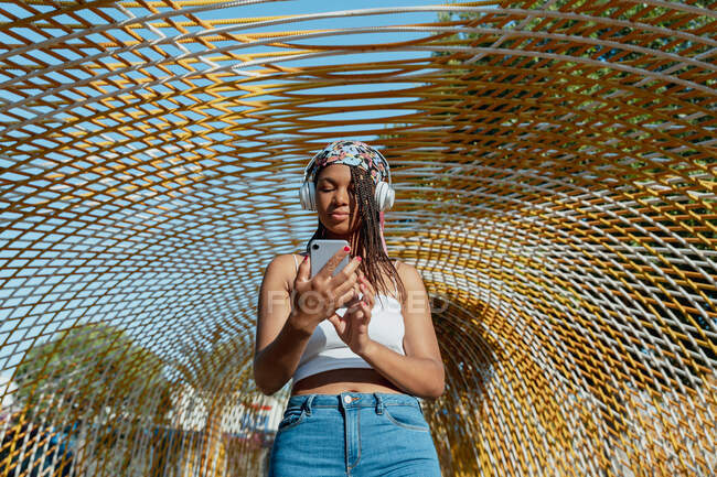 Модная молодая афроамериканка в беспроводных наушниках слушает музыку во время разговора по мобильному телефону на мосту — стоковое фото