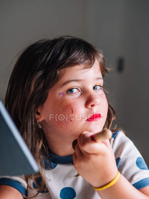 Kid with applicator making up face with assorted cosmetic products in house looking at camera — Stock Photo