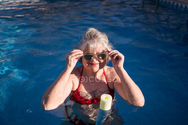 Femme âgée joyeuse avec des cheveux gris nageant dans la piscine et souriant brillamment à la caméra avec des lunettes de soleil — Photo de stock