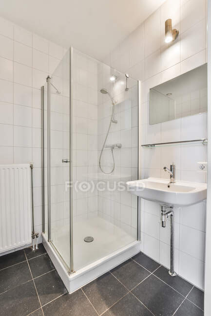 Интерьер домашней ванной комнаты с зеркалом, висящим над умывальником, размещенным рядом с входной дверью и стеклянной душевой кабиной в современной квартире — стоковое фото