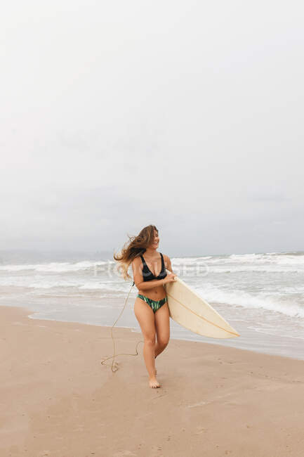 Cheerful young sportswoman in swimwear with surfboard looking away on sandy coast against stormy ocean — Stock Photo