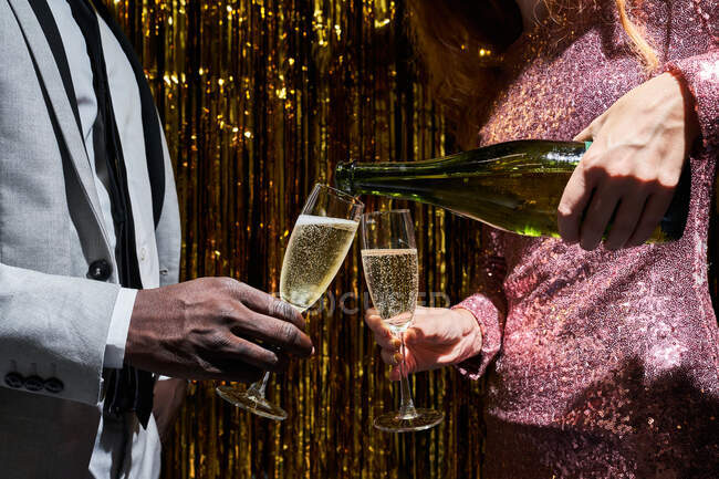 Crop anonymous woman pouring champagne from bottle into glass of black boyfriend during New Years Eve celebration against tinsel — Stock Photo
