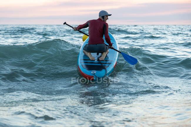 Vista posterior de surfista masculino irreconocible en traje de neopreno y sombrero en tabla de paddle surf en la orilla del mar - foto de stock