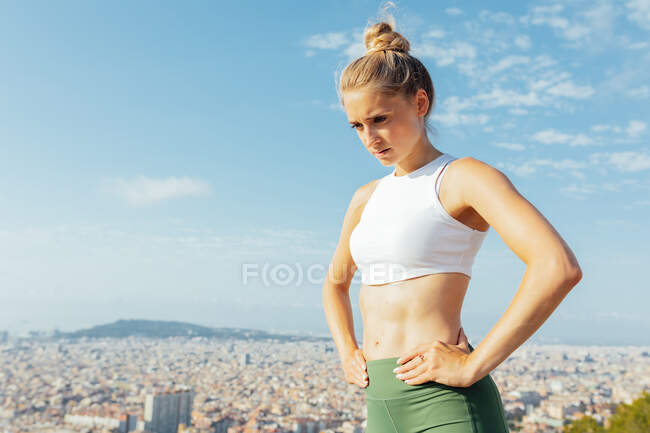 Продумано підійде жінка-спортсменка з руками на талії, милуючись літнім містом на сонячному світлі — стокове фото
