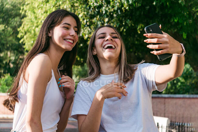 Alegre adolescente cerca del mejor amigo con acné tomando autorretrato en el teléfono celular a la luz del sol - foto de stock