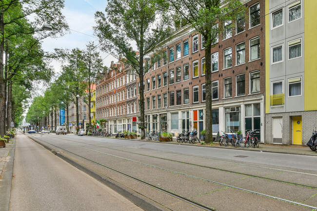 Envejecido exterior de la casa contra la carretera y bicicletas aparcadas entre árboles cubiertos durante el día en Amsterdam Holanda - foto de stock
