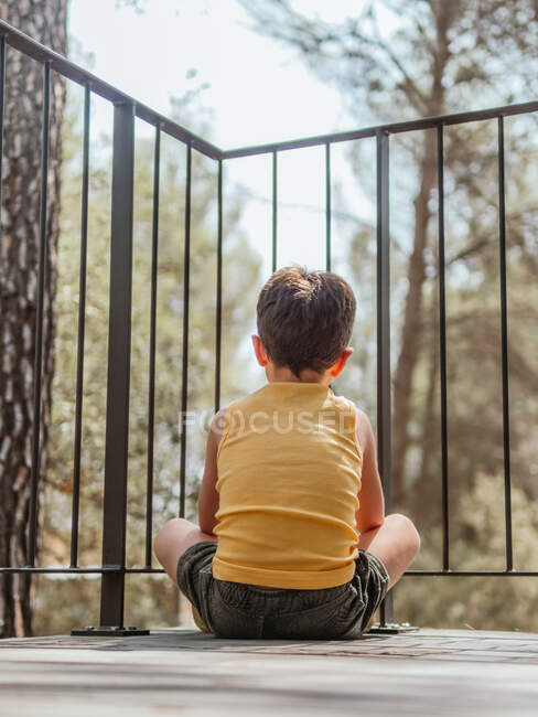 Vista trasera del niño sentado en la terraza de la casa de campo moderna situada en los bosques en verano - foto de stock
