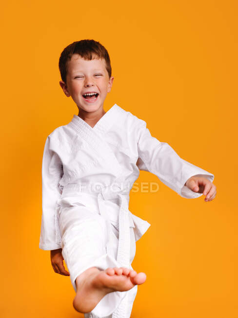 Front view of a smiling child dressed in a white kimono with eyes half closed and raising the right leg and foot in a studio session with orange background — Stock Photo