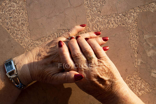 Crop unrecognizable elderly woman reaching out hands with red manicure and golden ring against beige wall in sunlight — Stock Photo