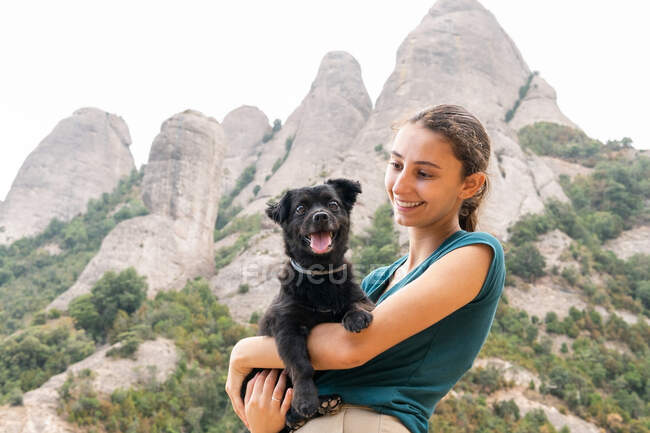 Zufriedene junge Touristin umarmt niedlichen Welpen mit Zunge gegen Montserrat mit Bäumen in Spanien — Stockfoto