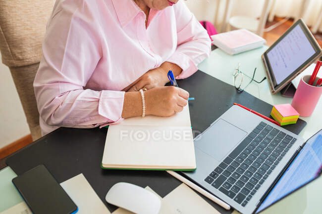 High angle of crop female entrepreneur taking notes in agenda while working at desk with netbook in workspace — Stock Photo