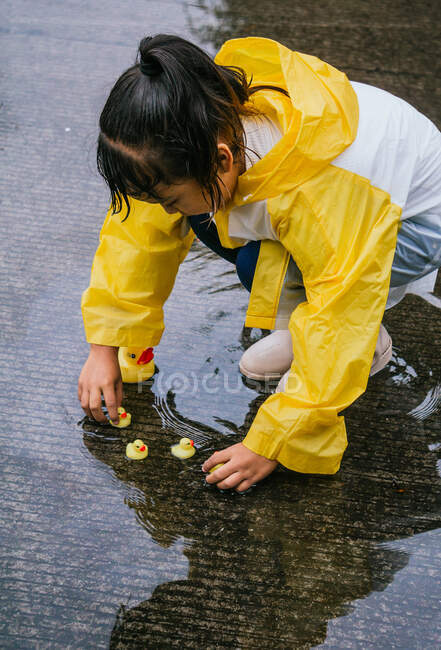 Charming ethnic kid in raincoat playing with plastic ducks reflecting in rippled puddle in rainy weather — Stock Photo
