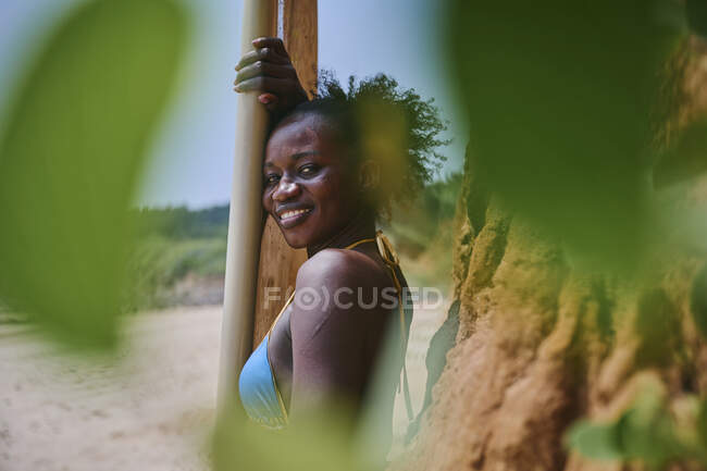 Vista lateral de la atleta afroamericana mirando a la cámara con tabla de surf desde un área de la playa enmarcada con plantas fuera de foco - foto de stock