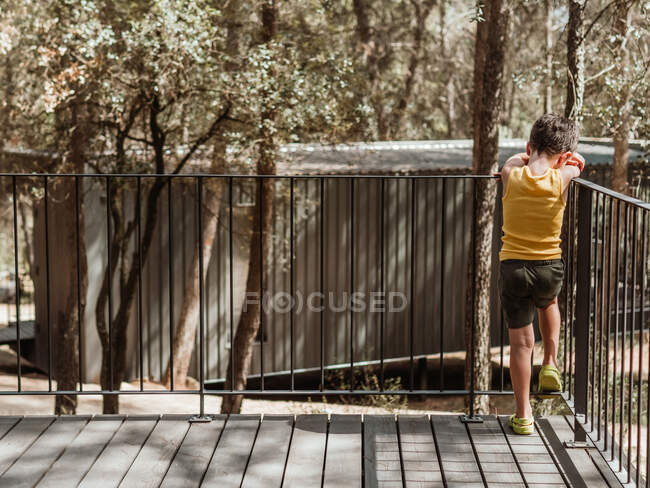 Rückansicht eines unkenntlichen kleinen Jungen, der im Sommer auf der Veranda eines modernen Ferienhauses im Wald steht, während er traurig weint — Stockfoto