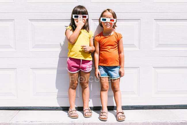 Filles mignonnes en vêtements colorés décontractés et lunettes tridimensionnelles debout sur fond blanc mur regardant la caméra — Photo de stock