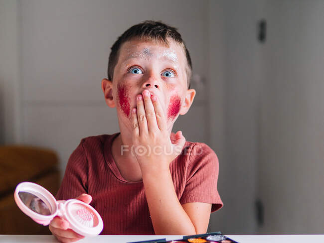 Amazed child with dirty face and mirror covering mouth while looking forward at table with eyeshadow palette in house — Stock Photo