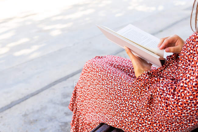 Zugeschnittene unkenntlich gemachte Frau in stilvoller Kleidung sitzt mit aufgeschlagenem Buch auf Holzbank gegen Gebäude mit Lichtwand am Tag — Stockfoto