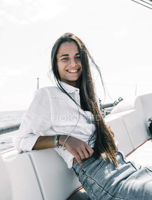 Созерцательная счастливая девушка-подросток, сидящая со скрещенными ногами на скамейке моторной лодки в океане, глядя в камеру на Тенерифе, Испания — стоковое фото