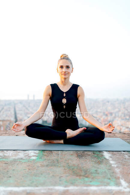 Full body of slim barefoot female in sportswear sitting in Ardha Padmasana with zen gesture during meditation on rooftop in evening — Stock Photo