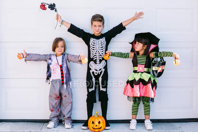 Pequeños amigos alegres en varios disfraces de Halloween con calabaza y accesorios levantando los brazos y mirando a la cámara mientras están de pie juntos cerca de la pared blanca - foto de stock