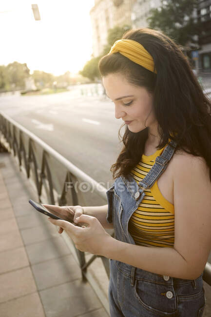 Visão lateral de jovem fêmea séria com cabelos longos em pé perto de cerca na calçada e navegação telefone móvel — Fotografia de Stock