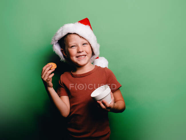 Adorable petit garçon souriant avec chapeau de Noël Père Noël prendre cookie de tasse sur fond vert en regardant la caméra — Photo de stock