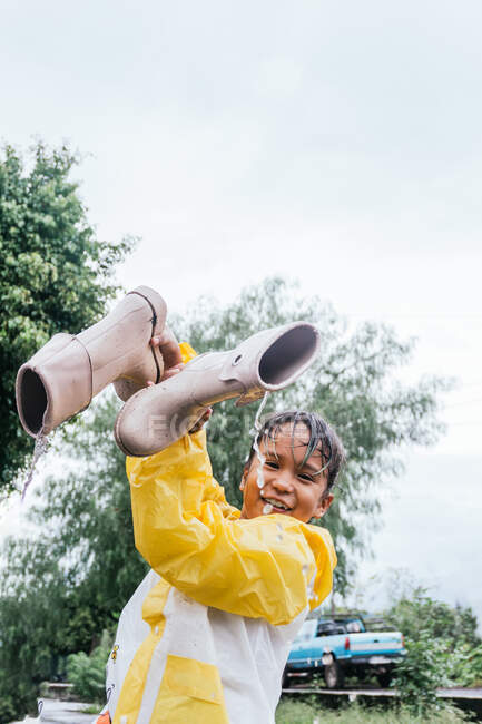 Smiling ethnic kid in slicker pouring aqua from gumboots while looking at camera in rainy weather — Stock Photo